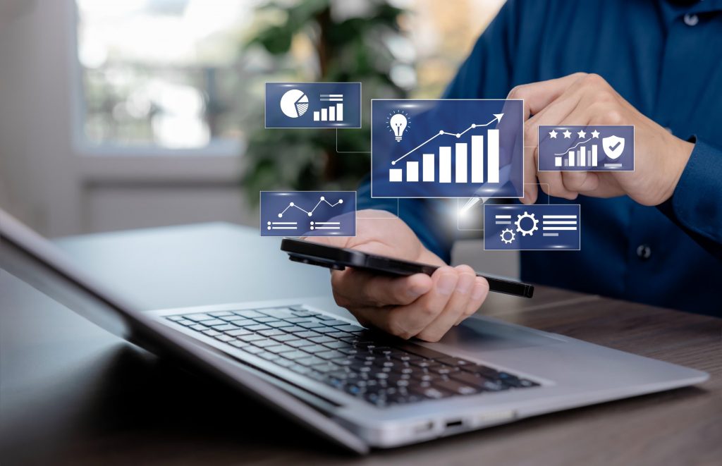 Executivo segurando um smartphone que projeta gráficos flutuantes de crescimento, engrenagens e dashboards, com um notebook na mesa, simbolizando a análise de dados na gestão da tecnologia da informação.