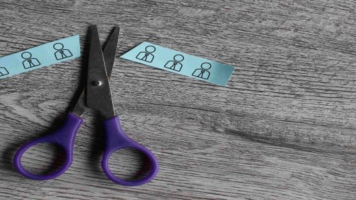 Tesoura cortando uma tira de papel com ícones de funcionários, simbolizando o corte de pessoal e a redução de equipe durante um layoff.