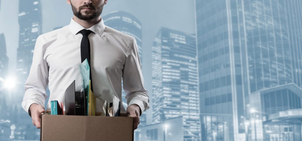 Homem de camisa e gravata segurando uma caixa de papelão com pertences de escritório após um processo de layoff, com prédios corporativos ao fundo.