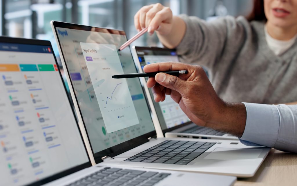 Pessoas apontando canetas digitais para telas de notebooks exibindo gráficos financeiros de gestão empresarial