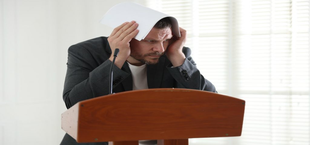 Homem ansioso se escondendo com papéis no púlpito, ilustrando o medo de falar em público.