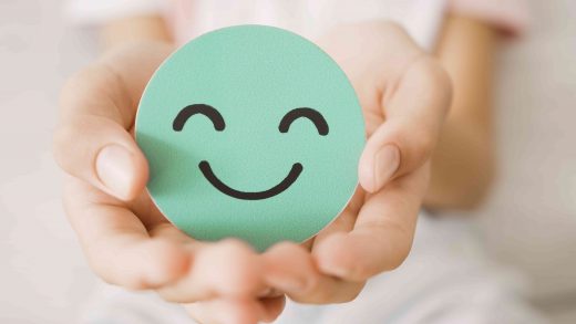Mãos segurando um círculo verde com um rosto sorridente desenhado, simbolizando o bem-estar, o equilíbrio emocional e a importância da saúde mental