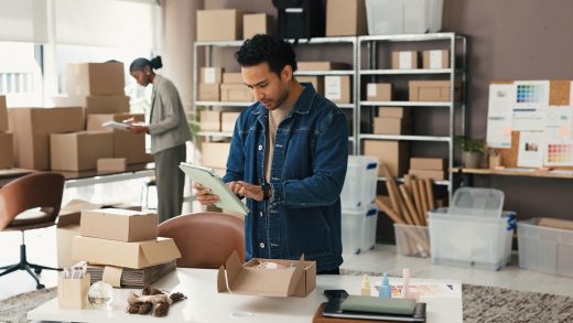 Analista de logística utilizando um tablet para gerenciar e organizar pedidos em mesas com várias caixas de papelão em um ambiente de trabalho de e-commerce