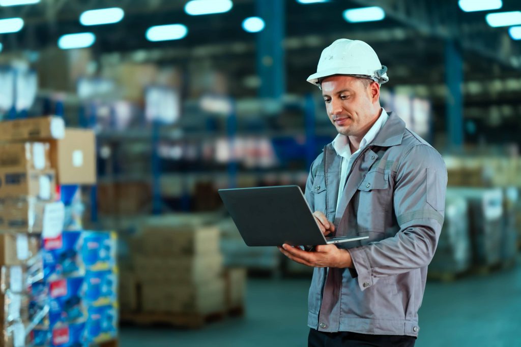 Profissional de logística usando capacete branco e uniforme, segurando um notebook enquanto confere dados em um grande armazém de distribuição com prateleiras e caixas ao fundo.